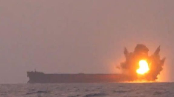 A ship on fire at sea, with an explosion visible on the vessel.